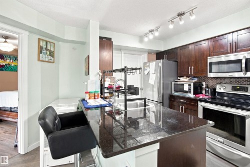 Kitchen featuring dark wood-finish cabinetry, stainless steel appliances, a dark stone countertop island, and track lighting - #413 18126 77 St Nw, Edmonton, AB 