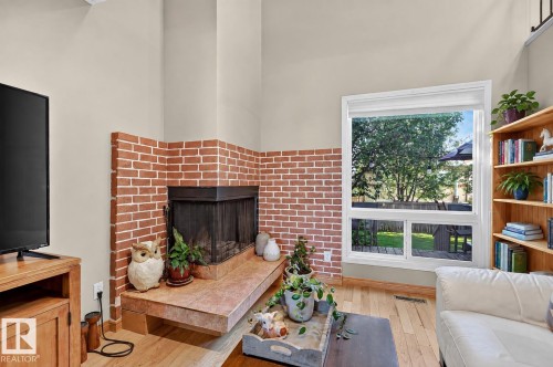 Brick-clad fireplace with a stone hearth, hardwood flooring, and a large window overlooking a green yard - 17919 78 Avenue Nw, Edmonton, AB 