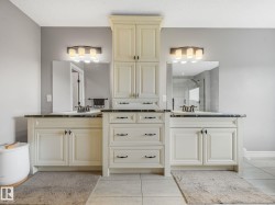 A luxurious primary ensuite bathroom featuring his-and-hers vanities, elegant cabinetry, and a spacious layout. Enjoy a deep soaking tub beside a large window, plus a generous glass-enclosed double shower — the perfect blend of comfort and elegance. - 