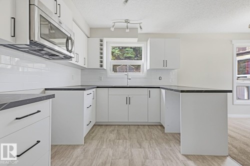 White cabinetry with matte black hardware, dark countertops, a subway tile backsplash, stainless steel microwave, and wood-finish flooring - 10621 Beaumaris Road Nw, Edmonton, AB 