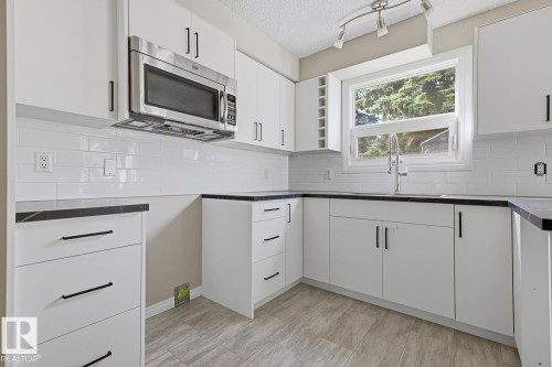 White cabinetry with black hardware, stainless steel microwave, white subway tile backsplash, dark countertops, and wood-finish flooring - 10621 Beaumaris Road Nw, Edmonton, AB 