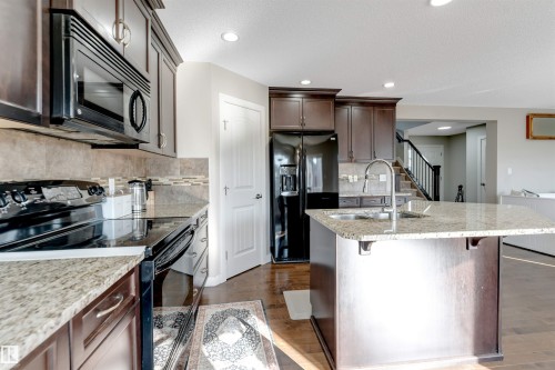 Kitchen featuring granite countertops, wood-finish flooring, dark wood cabinetry, a tile backsplash, and stainless steel appliances - 136 55 Street Sw, Edmonton, AB 