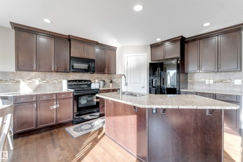 Kitchen featuring dark wood-finish cabinetry, light-toned countertops, black appliances, a central island with a sink, and recessed lighting - 136 55 Street Sw, Edmonton, AB 