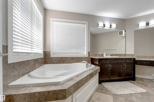 Built-in soaking tub with tile surround, dual vanity with dark wood cabinetry, integrated sink, and wall-mounted lighting fixtures - 136 55 Street Sw, Edmonton, AB 