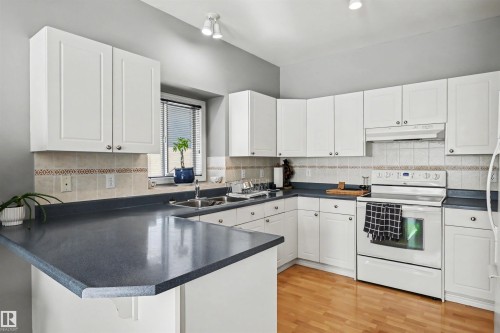 Kitchen featuring white cabinetry, dark countertops, a tiled backsplash, wood-finish flooring, and a built-in stainless steel sink - 8513 156 Avenue Nw, Edmonton, AB 