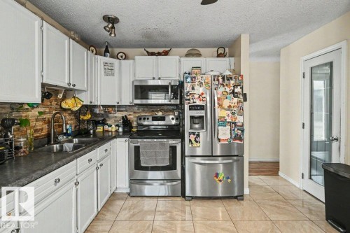 Kitchen featuring white cabinetry, stainless steel appliances, a stone tile backsplash, a double basin sink, and tile flooring - 8708 25A Avenue Nw, Edmonton, AB 