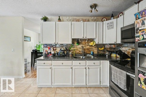 White cabinetry, stacked stone-finish backsplash, dark countertops, tile flooring, and integrated stainless steel appliances - 8708 25A Avenue Nw, Edmonton, AB 