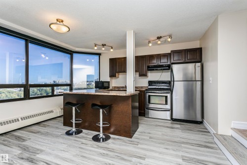 Expansive windows providing abundant natural light, wood-finish flooring, dark wood cabinetry, a built-in kitchen island, and stainless steel appliances - #815 10160 114 St Nw, Edmonton, AB 