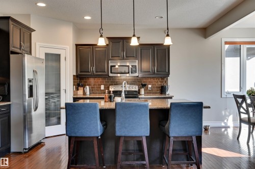 Kitchen featuring dark wood-finish cabinetry, stainless steel appliances, a tile backsplash, an island with countertop seating, and wood-finish flooring - 2203 Warry Loop Sw, Edmonton, AB 