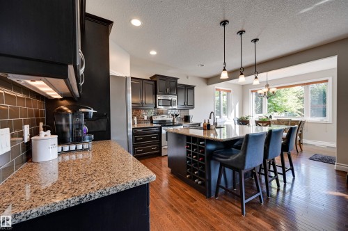 Kitchen featuring dark cabinetry, granite countertops, hardwood flooring, a central island with integrated wine storage, and recessed lighting - 2203 Warry Loop Sw, Edmonton, AB 