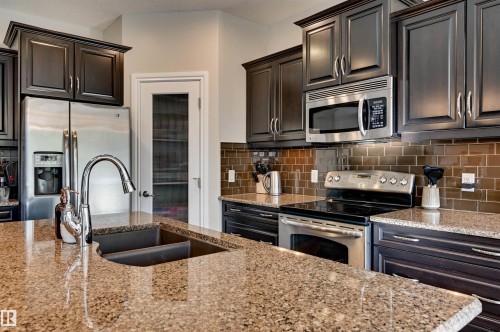 Spacious kitchen featuring stone countertops, stainless steel appliances, dark wood cabinetry, a tile backsplash, and a double basin sink with a gooseneck faucet - 2203 Warry Loop Sw, Edmonton, AB 