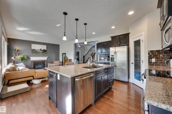 Open-concept living space with wood-finish flooring, a kitchen island with stone countertops, stainless steel appliances, and a gas fireplace with a stone surround - 