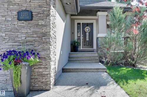 Stacked stone accent wall, concrete steps, dark paneled entry door with sidelights, porch with columns, and surrounding foliage - 2203 Warry Loop Sw, Edmonton, AB 