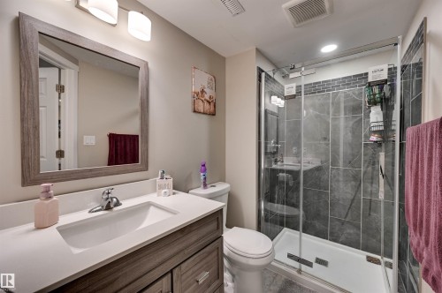 Glass-enclosed shower with dark tile surround, single vanity with integrated sink, wood-finish cabinetry, and framed mirror - 2203 Warry Loop Sw, Edmonton, AB 