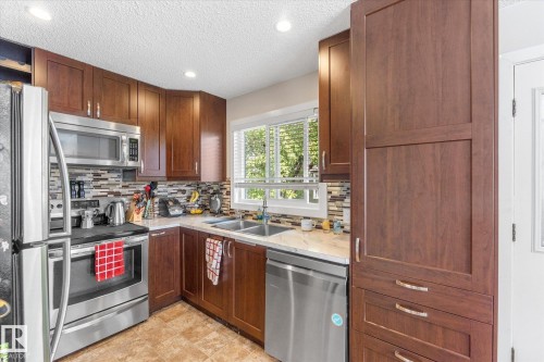 Kitchen featuring wood-finish cabinetry, stainless steel appliances, a mosaic tile backsplash, and light countertops - 16012 98 Avenue Nw, Edmonton, AB 