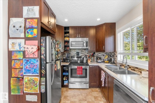 Kitchen featuring wood-finish cabinetry, stainless steel appliances, a mosaic tile backsplash, and a double basin sink - 16012 98 Avenue Nw, Edmonton, AB 