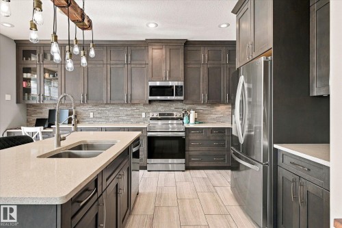 Kitchen featuring dark wood-finish cabinetry, stainless steel appliances, a stone tile backsplash, a center island with an undermount sink, and light-toned tile flooring - 2011 Genesis Lane, Stony Plain, AB 