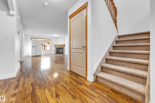 Hardwood-finish flooring extending from the entryway into the living area, featuring a carpeted staircase with a wood-finish banister, a white fireplace with a wood mantle, and white interior doors with wood trim - 10621 175A Avenue Nw, Edmonton, AB 