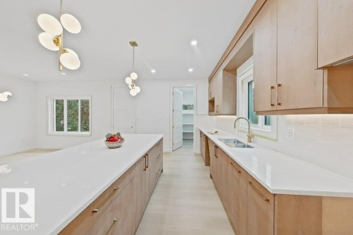 Spacious kitchen featuring a long center island with a light-toned countertop, wood-finish cabinetry with brass hardware, and a double-basin sink with a gold-tone faucet - 9216 91 Street Nw, Edmonton, AB - Indoor Photo Showing Kitchen With Double Sink