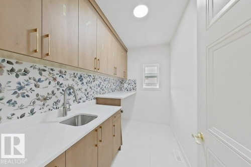 Light-filled utility space featuring extensive light wood-finish cabinetry with gold-tone hardware, a floral-patterned backsplash, white countertops, and an undermount sink with a gooseneck faucet - 9216 91 Street Nw, Edmonton, AB - Indoor Photo Showing Kitchen