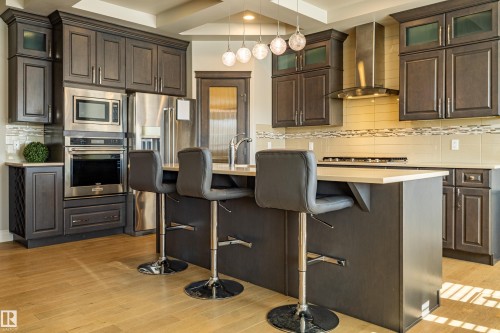 Kitchen featuring dark wood cabinetry, stainless steel appliances, light-toned countertops, a central island with bar seating, and wood-finish flooring - 233 Hawks Ridge Boulevard Nw, Edmonton, AB 