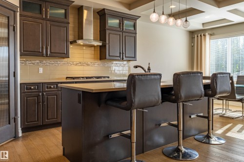 Dark wood cabinetry with glass-front accents, stainless steel range hood, light wood-finish flooring, and a multi-pendant light fixture - 233 Hawks Ridge Boulevard Nw, Edmonton, AB 