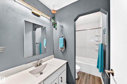 Vanity with integrated sink, modern wall-mounted light fixture, full-width mirror, and a shower-tub combination with wood-finish flooring - 5620 93A Avenue Nw, Edmonton, AB 