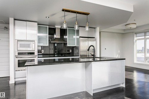 Modern kitchen featuring an integrated stainless steel appliance suite, black countertop island with sink, white high-gloss cabinetry, and pendant lighting - #1902 10388 105 St Nw, Edmonton, AB 