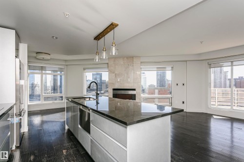 Open-concept living space featuring dark wood-finish flooring, a kitchen island with an undermount sink and dark countertop, a built-in fireplace with a stone-finish surround, and expansive windows - #1902 10388 105 St Nw, Edmonton, AB 