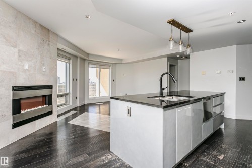 Contemporary kitchen island with a dark countertop and integrated sink, pendant lighting fixture, dark wood-finish flooring, and a fireplace with a stone-finish surround - #1902 10388 105 St Nw, Edmonton, AB 
