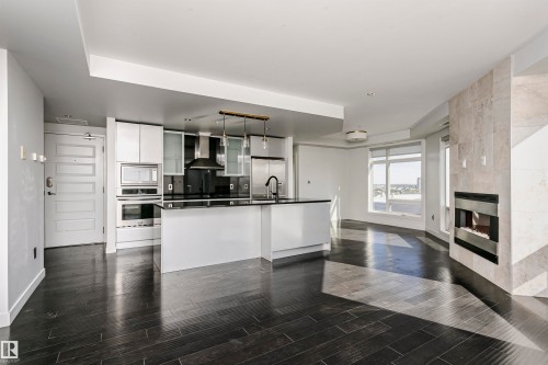 Open-concept living space featuring wood-finish flooring, a modern kitchen island with an undermount sink, stainless steel appliances, and a built-in fireplace with a stone surround - #1902 10388 105 St Nw, Edmonton, AB 
