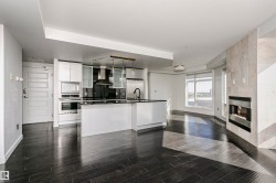 Open-concept living space featuring wood-finish flooring, a modern kitchen island with an undermount sink, stainless steel appliances, and a built-in fireplace with a stone surround - 
