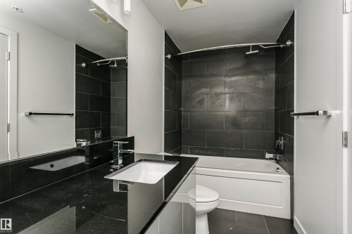Bathroom featuring a built-in tub with a tile surround, dark tile flooring, a large vanity mirror, and a sleek vessel sink with chrome fixtures - #1902 10388 105 St Nw, Edmonton, AB 