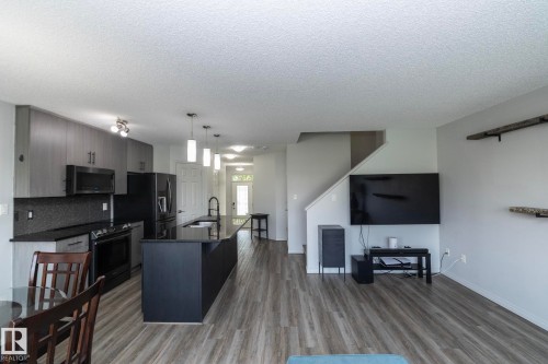Open-concept living space featuring wood-finish flooring, a modern kitchen with dark cabinetry and stainless steel appliances, a kitchen island with pendant lighting, a dark backsplash, and a staircase - 1640 Chapman Way Sw, Edmonton, AB - Indoor Photo Showing Kitchen