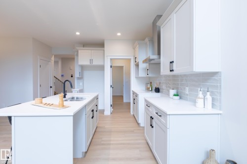 Modern kitchen featuring white cabinetry with black hardware, light wood-finish flooring, a center island with an undermount sink, and a stainless steel range hood - 6306 65 Ave, Beaumont, AB 