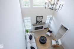 Two-story ceiling, multiple windows, fireplace with white brick surround and wood-finish mantel, wood-finish flooring, and a contemporary black chandelier - 