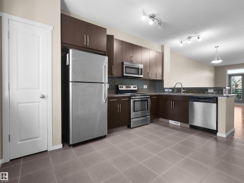 Kitchen featuring stainless steel appliances, dark wood-finish cabinetry, tile flooring, and track lighting - #162 603 Watt Bv Sw, Edmonton, AB 