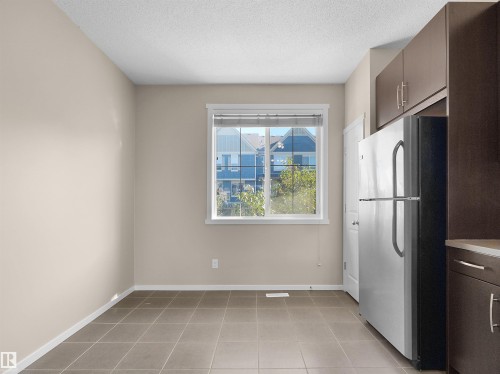 Stainless steel refrigerator, wood-finish cabinetry, neutral tile flooring, window with white trim, and white paneled door - #162 603 Watt Bv Sw, Edmonton, AB 