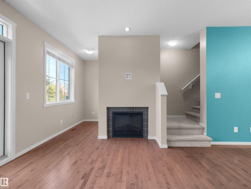Hardwood-style flooring throughout, featuring a fireplace with dark tile surround, large windows, and a carpeted staircase - #162 603 Watt Bv Sw, Edmonton, AB 