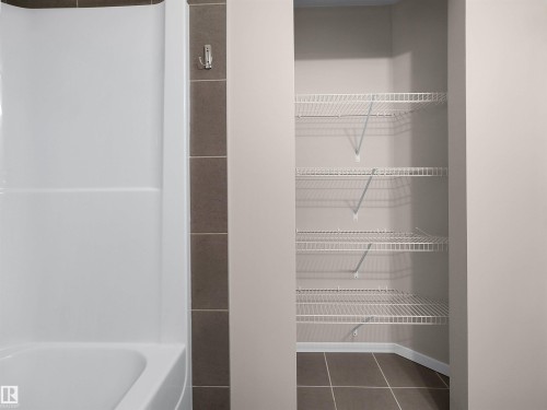 White bathtub with integrated shower, brown vertical tile accent, and a built-in closet with wire shelving and tile flooring - #162 603 Watt Bv Sw, Edmonton, AB 