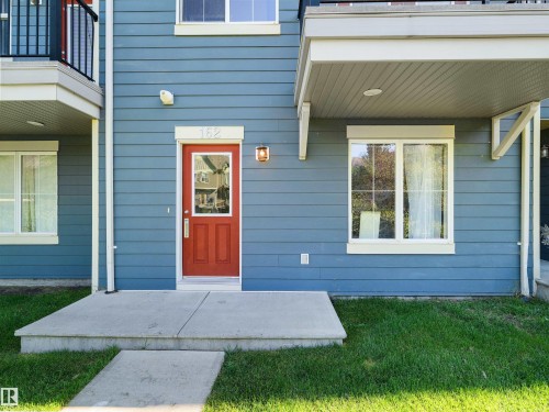 Blue horizontal siding exterior with a red paneled entry door, concrete entry pad, and white window trim - #162 603 Watt Bv Sw, Edmonton, AB 