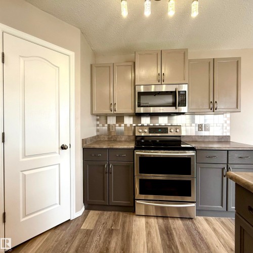Wood-finish flooring, two-tone cabinetry with upper and lower storage, stainless steel electric range with double ovens, stainless steel microwave, and tile backsplash - 910 Oakland Boulevard, Devon, AB 
