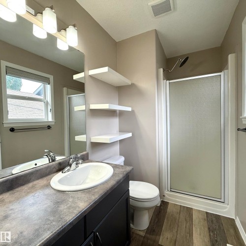 Bathroom featuring a vanity with an integrated sink, a large wall-mounted mirror, wood-finish flooring, a shower enclosure with a frosted glass door, and white floating shelves - 910 Oakland Boulevard, Devon, AB 