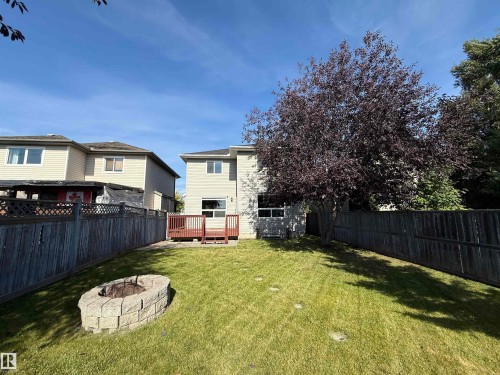 Expansive backyard featuring a manicured lawn, a built-in stone fire pit, a wooden privacy fence, a mature tree, and a wooden deck - 910 Oakland Boulevard, Devon, AB 