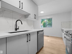 Modern kitchen featuring light wood-finish flooring, white cabinetry with matte black hardware, stone countertops, a tiled backsplash, and a stainless steel dishwasher - 