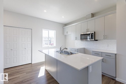 Kitchen featuring an island with a dual-basin stainless steel sink, light grey cabinetry, white subway tile backsplash, and wood-finish flooring - 3611 Triomphe Boulevard, Beaumont, AB 
