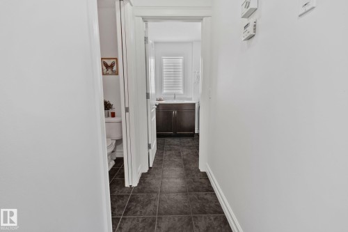 Hallway with dark tile flooring and white walls, leading to a bathroom with a dark wood-finish vanity and white window blinds - 1115 61 Street Sw, Edmonton, AB 