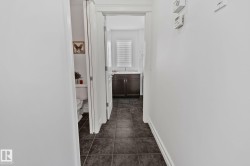 Hallway with dark tile flooring and white walls, leading to a bathroom with a dark wood-finish vanity and white window blinds - 
