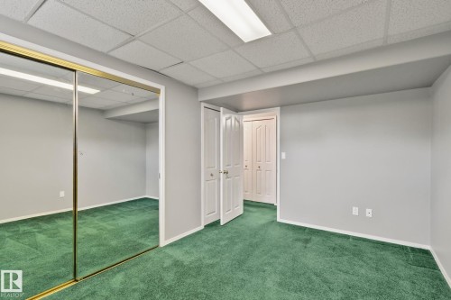 Green carpeting, full-wall mirrored closet with gold-toned trim, drop ceiling with fluorescent lighting, and white bi-fold doors - 14724 73 Street Nw, Edmonton, AB 