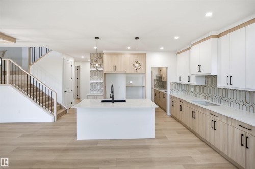 Gourmet kitchen featuring a central island, wood-finish flooring, full-height cabinetry, geometric tile backsplash, and a staircase with wood railing and black spindles - 13144 187 Avenue Nw, Edmonton, AB 